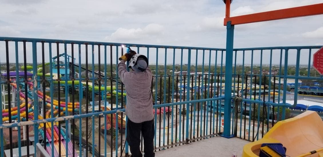 M&R Welding fabricator welding railing atop a ride tower at Hurricane Harbor, Arlington, TX, with the park and Arlington skyline in the background